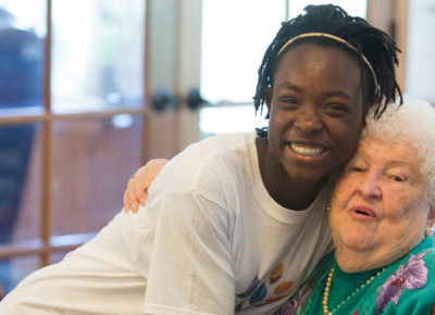 Young lady happily hugging an elderly women who is sitting down as she makes a difference by volunteering.