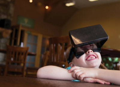 Smiling little girl sitting at a table wearing a virtual reality headset.