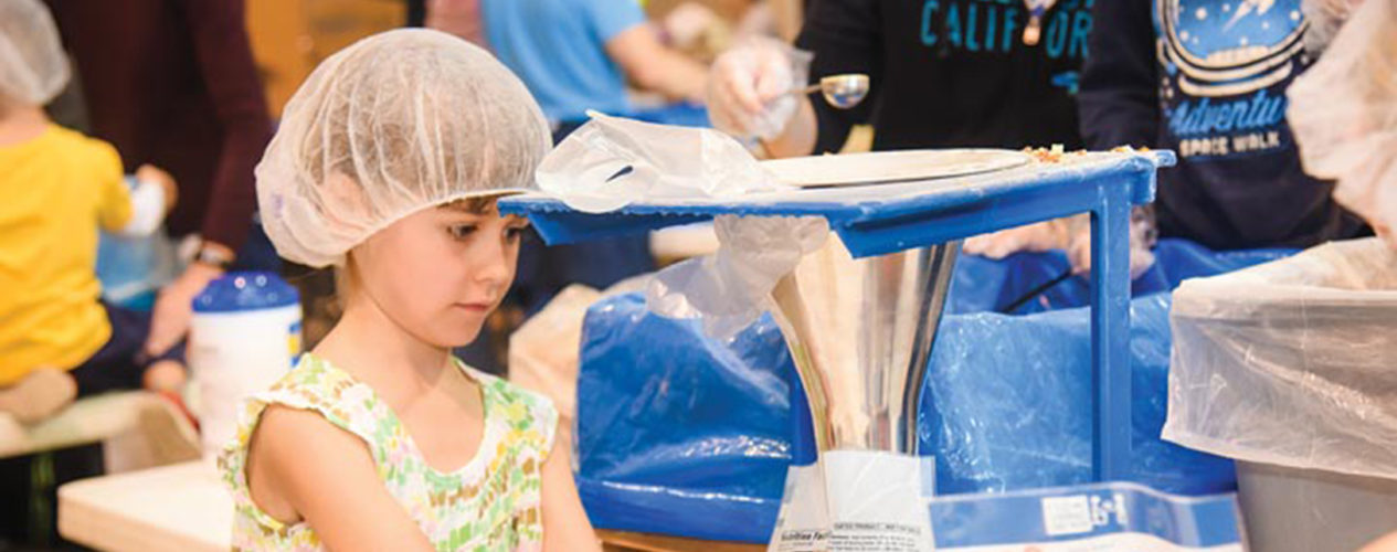 Young girl in a hair net is holding a bag under a funnel, waiting for a scoop of vitamins to be placed in the bag.