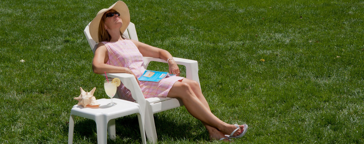 A woman is resting in a chair on a grassy lawn. She has on a sun hat and sun glasses. There is a book in her lap.