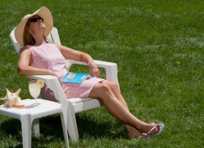 A woman is resting in a chair on a grassy lawn. She has on a sun hat and sun glasses. There is a book in her lap.