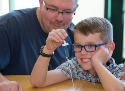 A son is holding a magnet, attracting a paper clip on a string, as his father watches.