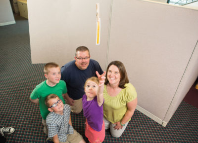 A family consisting of a mom, dad, two sons and a daughter are reaching for a measuring tape hanging from the ceiling. The taller family members are on their knees.