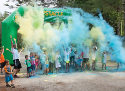 A group of kids huddle under a running finish line as powdered colored chalk shoots through the air.