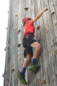 Middle school boy in a red shirt and red helmet scales a wooden rock climbing wall.