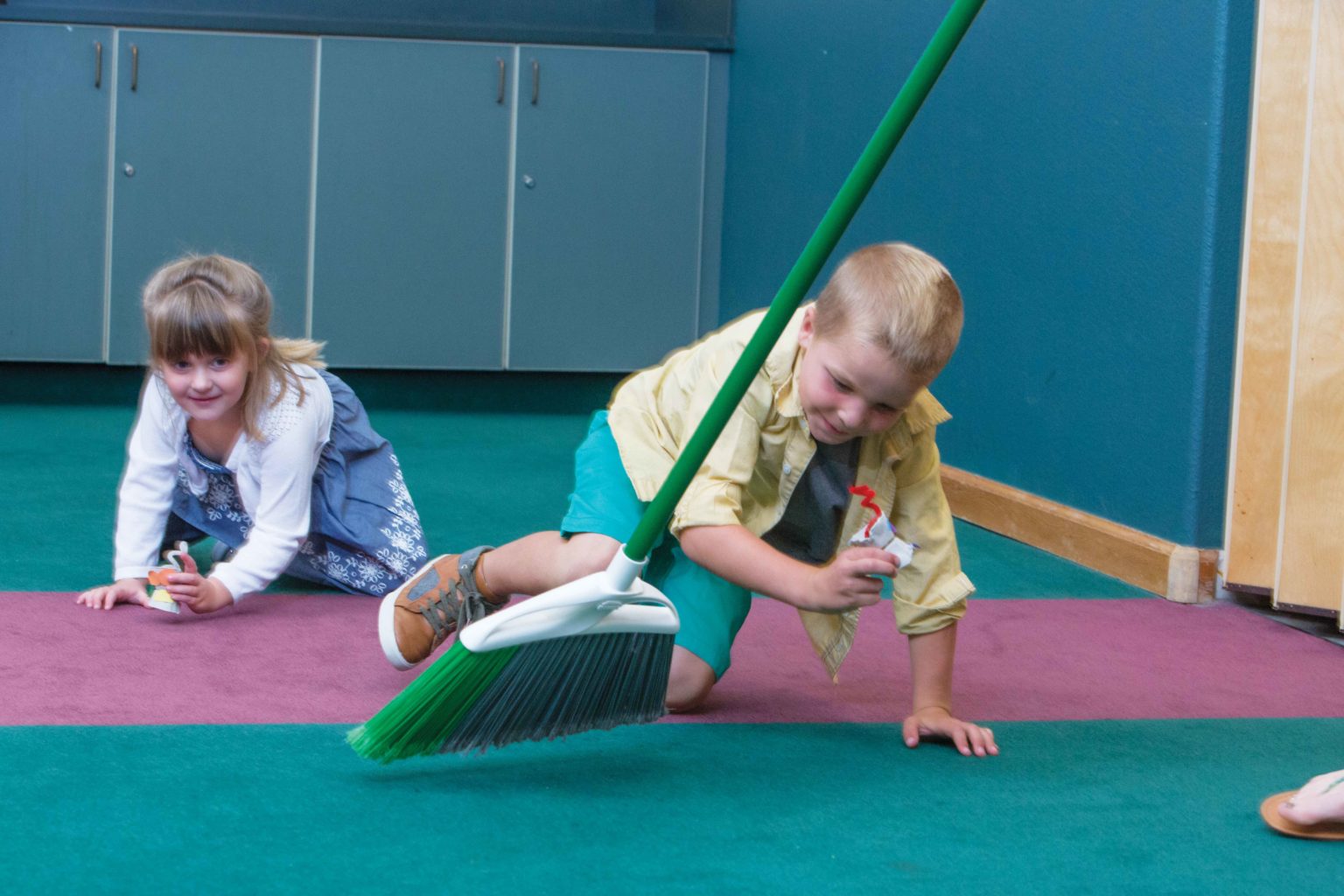 Two preschoolers crawling around protecting their craft robots from a broom.