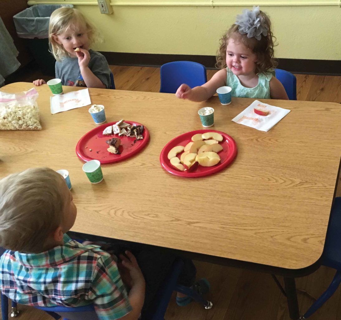 Three prechoolers enjoy snack at a table.