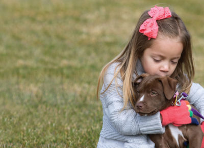 Elementary aged girl sitting outside in the grass, hugging a puppy. She's wearing a warm sweater and gloves as she gives the puppy a kiss on it's head.