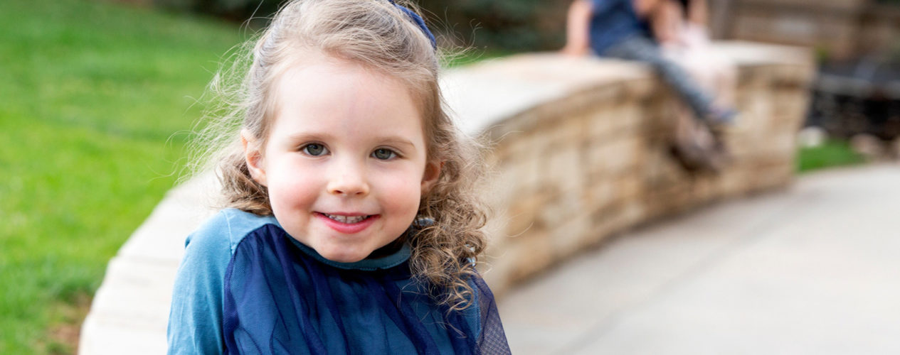 Little girl with curly hair smiles at the camera. She's sitting outside on a small stone retaining wall.