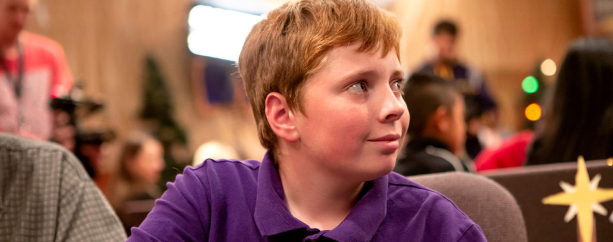 A preteen boy in a purple polo sits at a table, smiling.