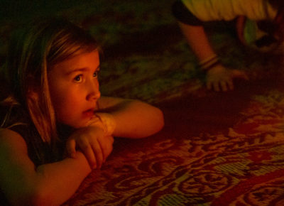 Preteen girl is thinking as she looks a a bright candle in a dark room. She is laying on her stomach with her head resting on her hands.