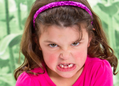 An elementary girl with read cheeks is sitting her bottom teeth out as she scowls at the camera.