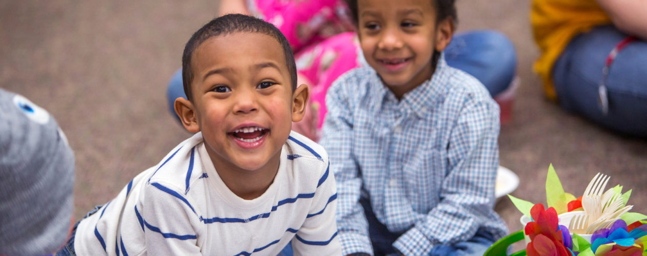 Two preschool boys smile brightly at the camera. They are sitting on their classroom floor.