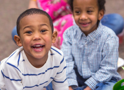 Two preschool boys smile brightly at the camera. They are sitting on their classroom floor.