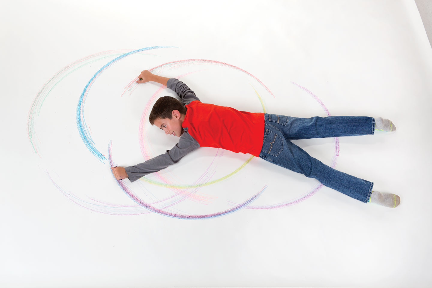 Boy laying on a white sheet of paper with crayons in his hands. He is laying on his stomach and moving his hands in an arch.