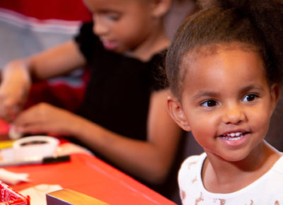 A preschool girl is smiling as she sits at a table covered in bows and wrapped presents.