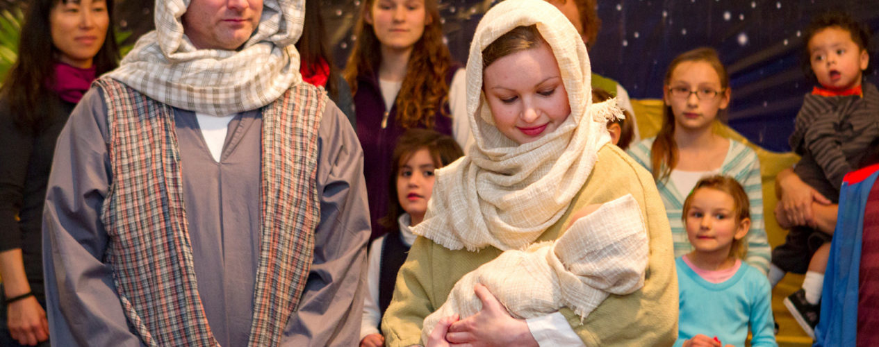 Two adults dressed up as Mary and Joseph are carrying a babydoll. There is a group of kids watching their skit.