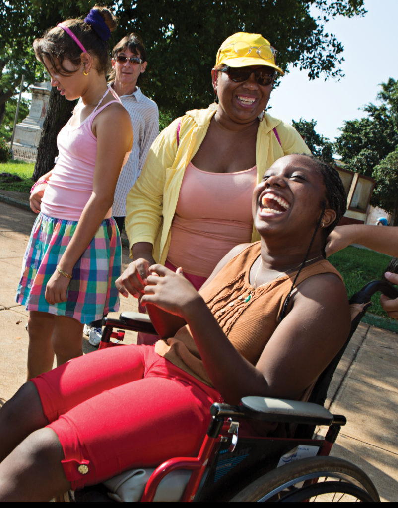 Teenage girl with Cerebral Palsy laughing with friends who are gathered around her wheelchair.