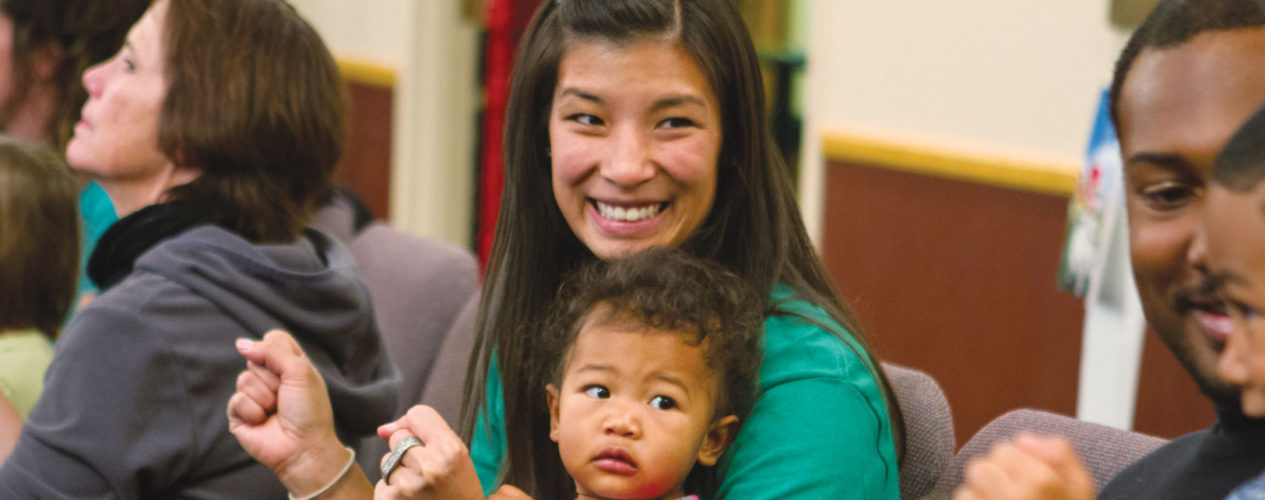 A mom smiles excitedly with a baby on her lap. She is looking towards her husband and preschool-age son.