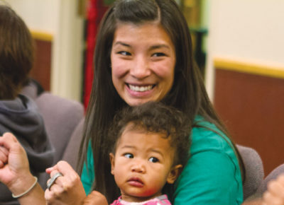 A mom smiles excitedly with a baby on her lap. She is looking towards her husband and preschool-age son.