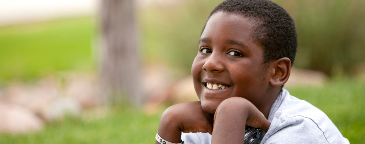 An older elementary-aged boy is sitting outside, smiling as he rests his chin on his hand.