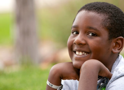 An older elementary-aged boy is sitting outside, smiling as he rests his chin on his hand.