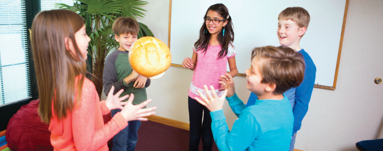 Five preteen children standing in a circle tossing around a mid-sized loaf of bread.