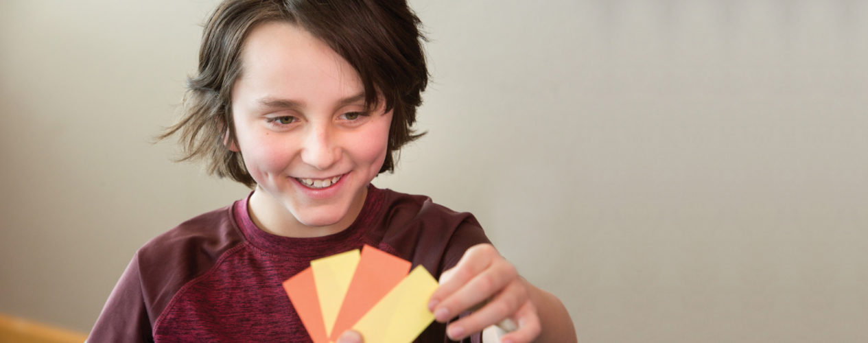 An older elementary boy standing in front of a beige wall. He's picking from four cards, two that are yellow and two that are orange. He's smiling.