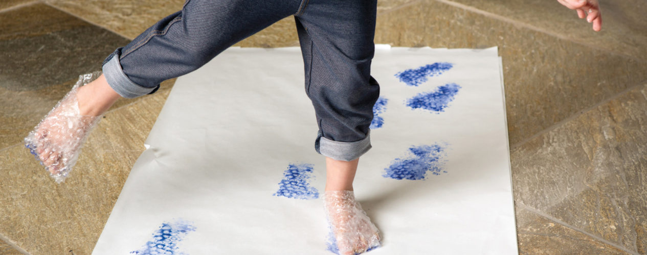 A preschool girl balances on one foot. She is wearing bubble wrap around both feet. The bubble wrap has been dipped in blue paint and she is walking across white paper.