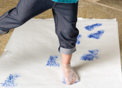 A preschool girl balances on one foot. She is wearing bubble wrap around both feet. The bubble wrap has been dipped in blue paint and she is walking across white paper.