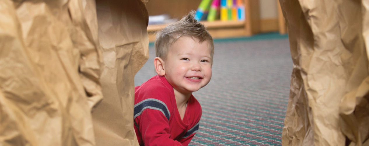 A toddler is crawling through a cave made out of brown paper bags. He is smiling as he participates in this nursery Bible activity.