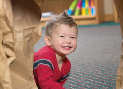 A toddler is crawling through a cave made out of brown paper bags. He is smiling as he participates in this nursery Bible activity.