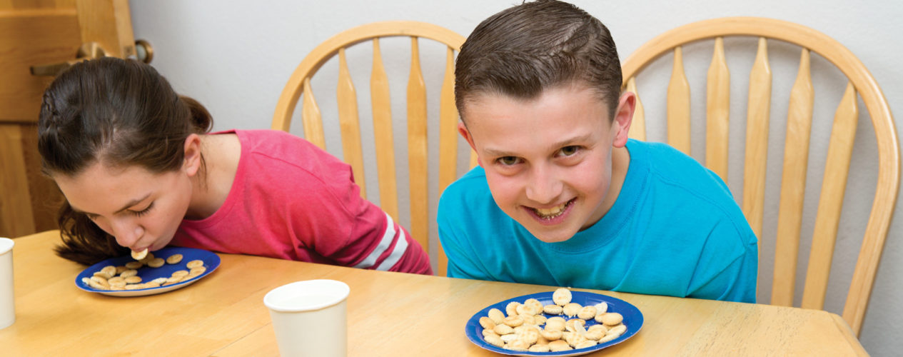 A preteen boy in a blue shirt and a preteen girl in a pink shirt eat oyster crackers of plates without using their hands.
