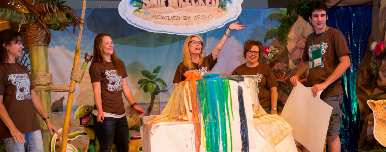 Five adults in VBS shirts standing on stage under "Shipwrecked VBS" logo. They've just completed a colorful demonstration and there's color overflowing from the demonstration table.