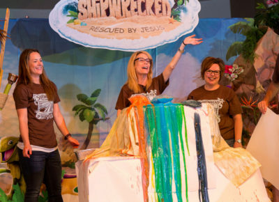 Five adults in VBS shirts standing on stage under "Shipwrecked VBS" logo. They've just completed a colorful demonstration and there's color overflowing from the demonstration table.