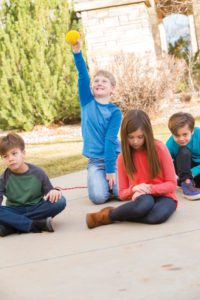 A group of children are sitting on the ground outside. Everyone looks sad, except one boy who is smiling as he holds a nerf ball up victoriously. 