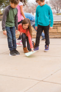 Children playing outside. They are placing colored construction paper on the ground and stepping on them.