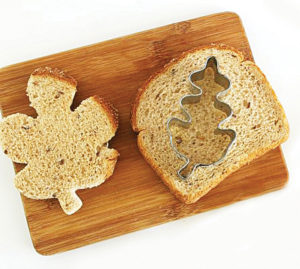 A piece of toast sitting on a cutting board. There's one piece of toast cut into the shape of a leaf, with a whole slice of bread next to it with a cutter on top.