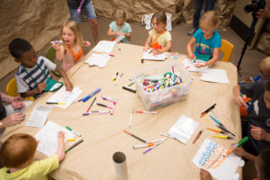 A group of children in early elementary school sit at a table covered in brown paper. They are coloring and there are markers scattered around.