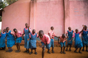 A group of children in their school uniforms singing and dancing outside their school.