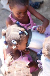 A preschool girl helping her younger sister drink from a tin cup.