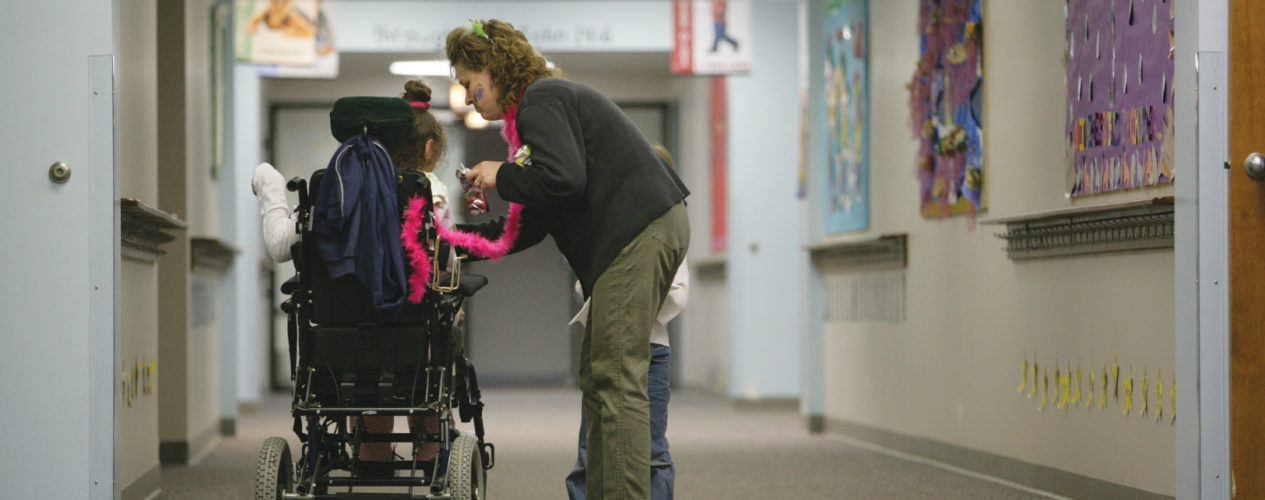 A mom stands in a hallway of a church helping her daughter who is in a wheel chair. Her son is standing next to her, as well.