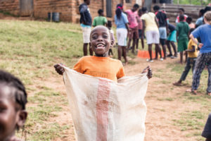 A child playing in a potato sack race as he smiles. There are a bunch of children outside playing.