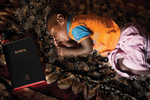 A preschool Ugandan child sleeping on carpet with a Bible next to him.