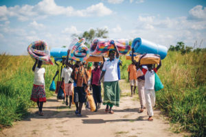 A group of women in Uganda carrying bedding on their heads as they walk through a field.