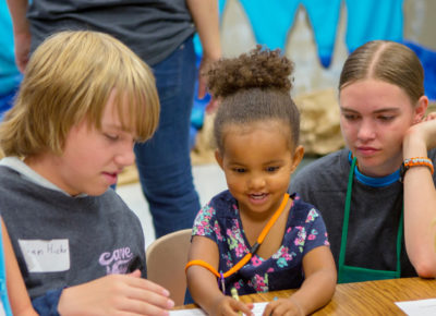 Two volunteers help a group of early elementary children with a craft.