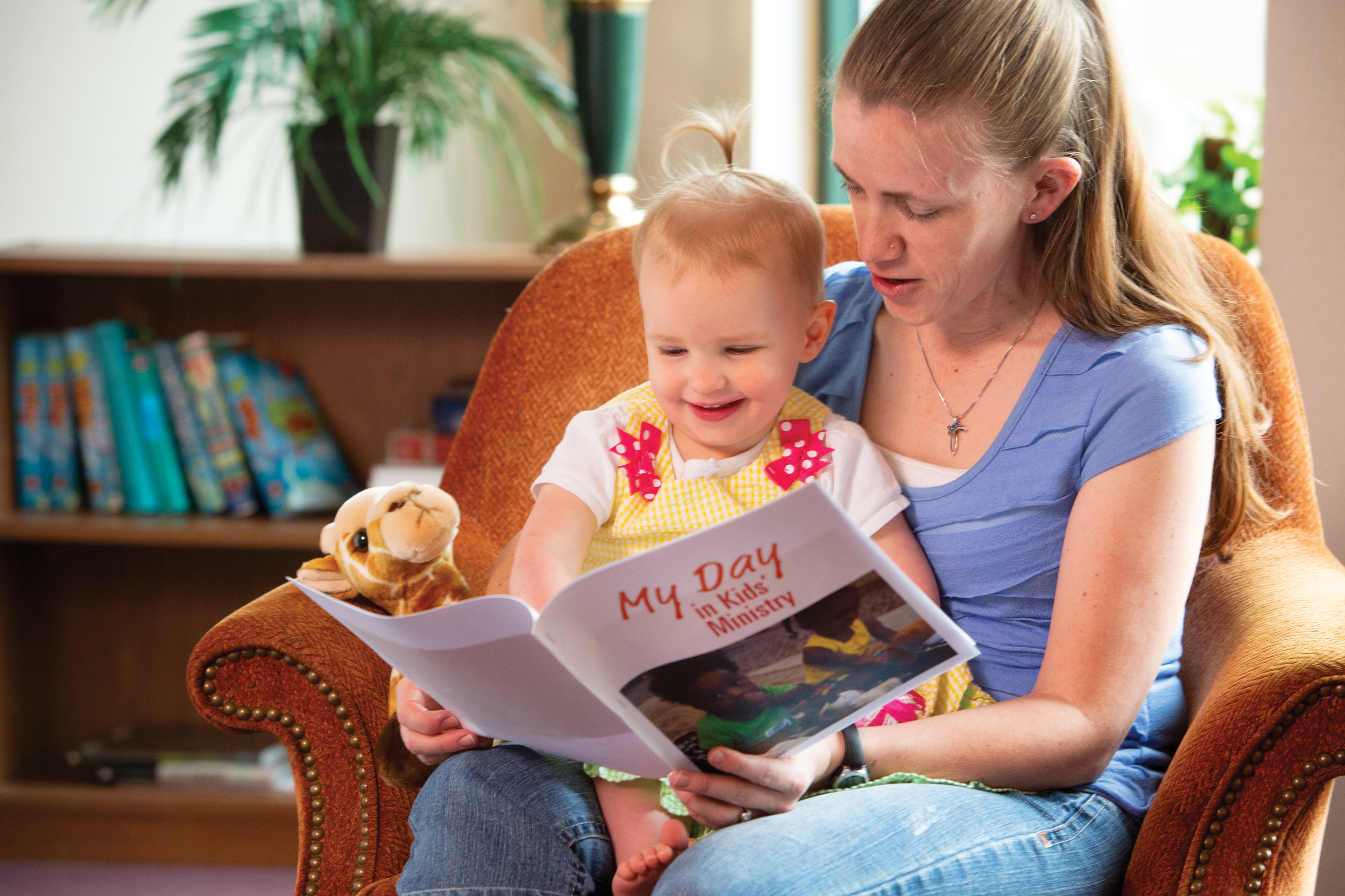 A mom is sitting in an arm chair with her two year old daughter on her lap. She is reading a "My Day in Kid's Ministry" book.