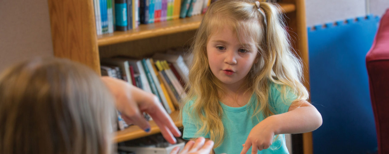 A female volunteer helps teach a preschool girl the Noah Ark finger play from this list of Bible activities. They are in front of a book case in a preschool children's ministry room.