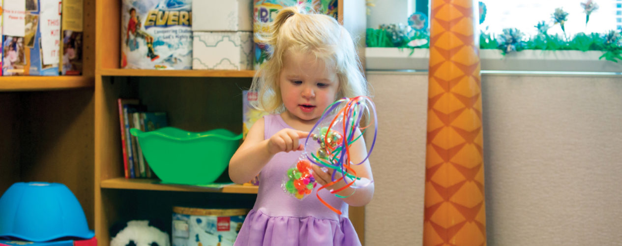 A toddler girl wearing a light purple dress is holding a celebration shaker in her hands.