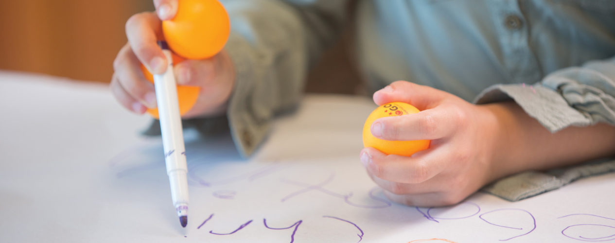An elementary-aged boy holding two orange ping pong balls in each hand. He's trying to write with a marker while he's holding the ping pong balls. This is one of the challenges in this list of Elementary Bible activities.